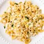 A white plate filled with bow tie lemon ricotta pasta, topped with creamy sauce, grated cheese, chopped herbs, and freshly ground black pepper.