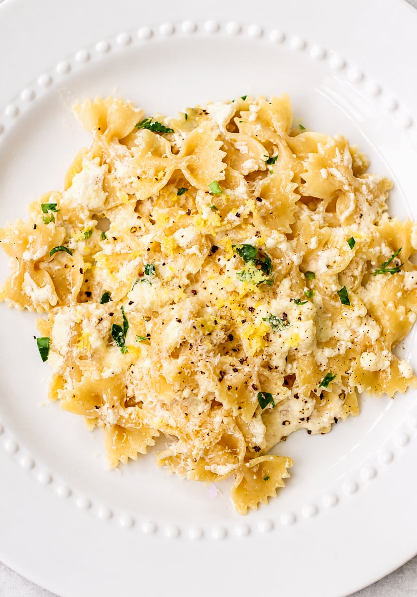 A white plate filled with bow tie lemon ricotta pasta, topped with creamy sauce, grated cheese, chopped herbs, and freshly ground black pepper.