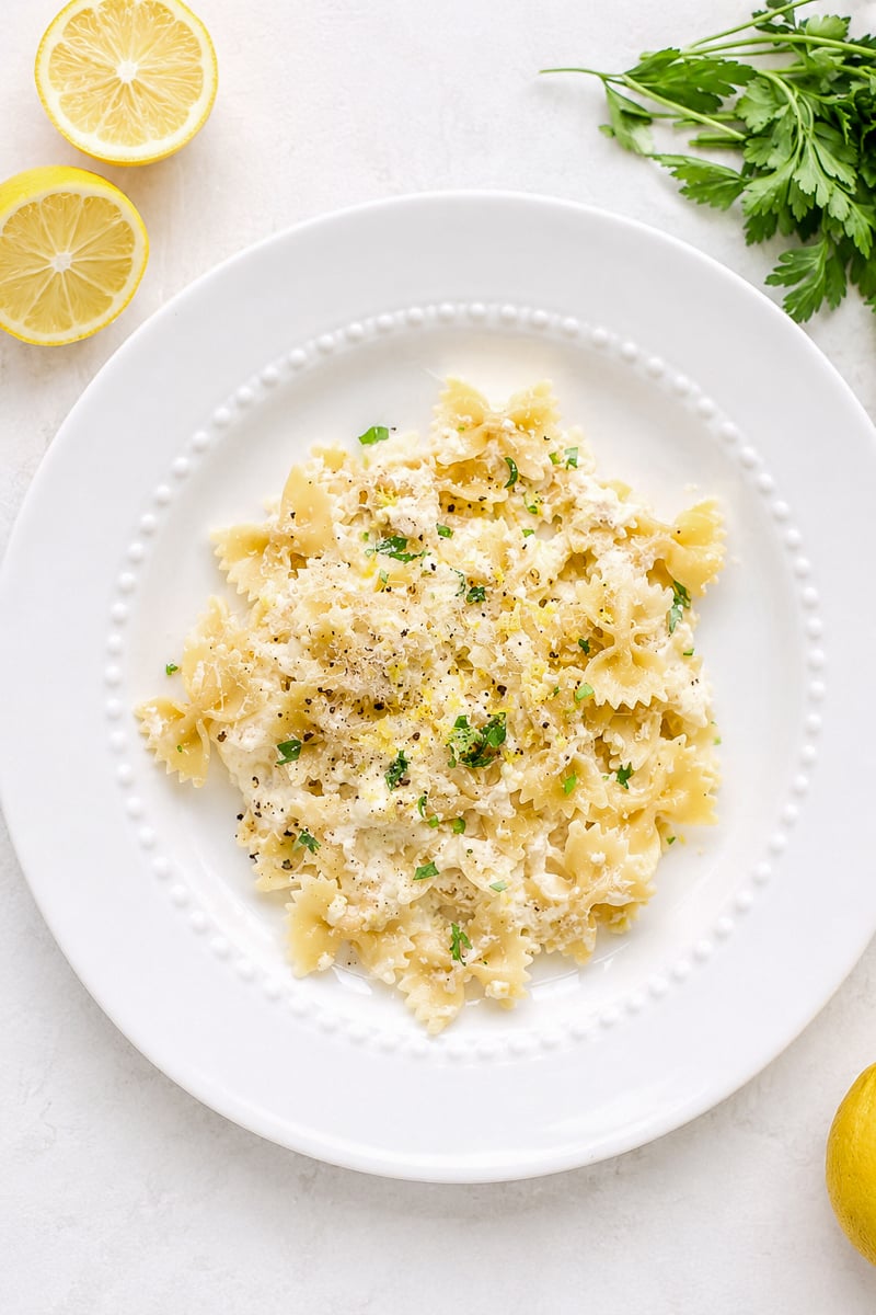 A white plate of lemon ricotta pasta topped with grated cheese, chopped parsley, and black pepper. Lemon halves and fresh parsley are arranged on the white surface beside the plate.
