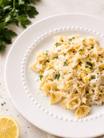 A white plate of lemon ricotta pasta in a creamy sauce, topped with chopped parsley, black pepper, and grated lemon zest. Fresh parsley and a lemon slice sit nearby on a light-colored surface.
