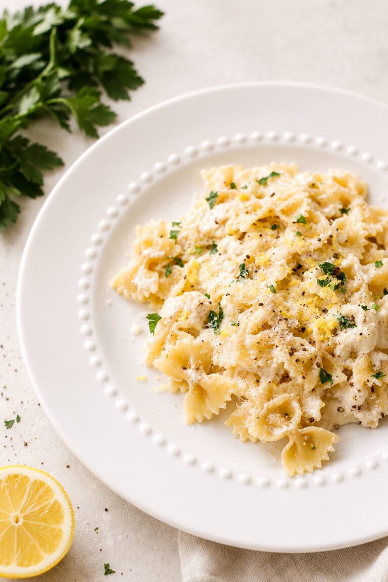 A white plate of lemon ricotta pasta in a creamy sauce, topped with chopped parsley, black pepper, and grated lemon zest. Fresh parsley and a lemon slice sit nearby on a light-colored surface.