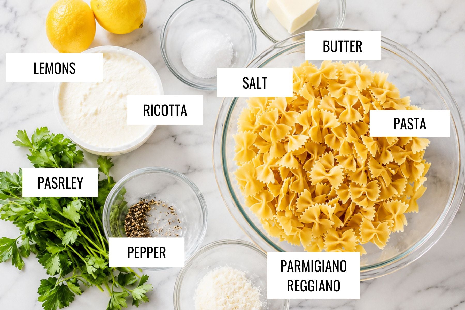 Bowls and ingredients on a marble surface for lemon ricotta pasta, labeled: lemons, ricotta cheese, parsley, pepper, salt, butter, uncooked bowtie pasta, and Parmigiano Reggiano cheese.