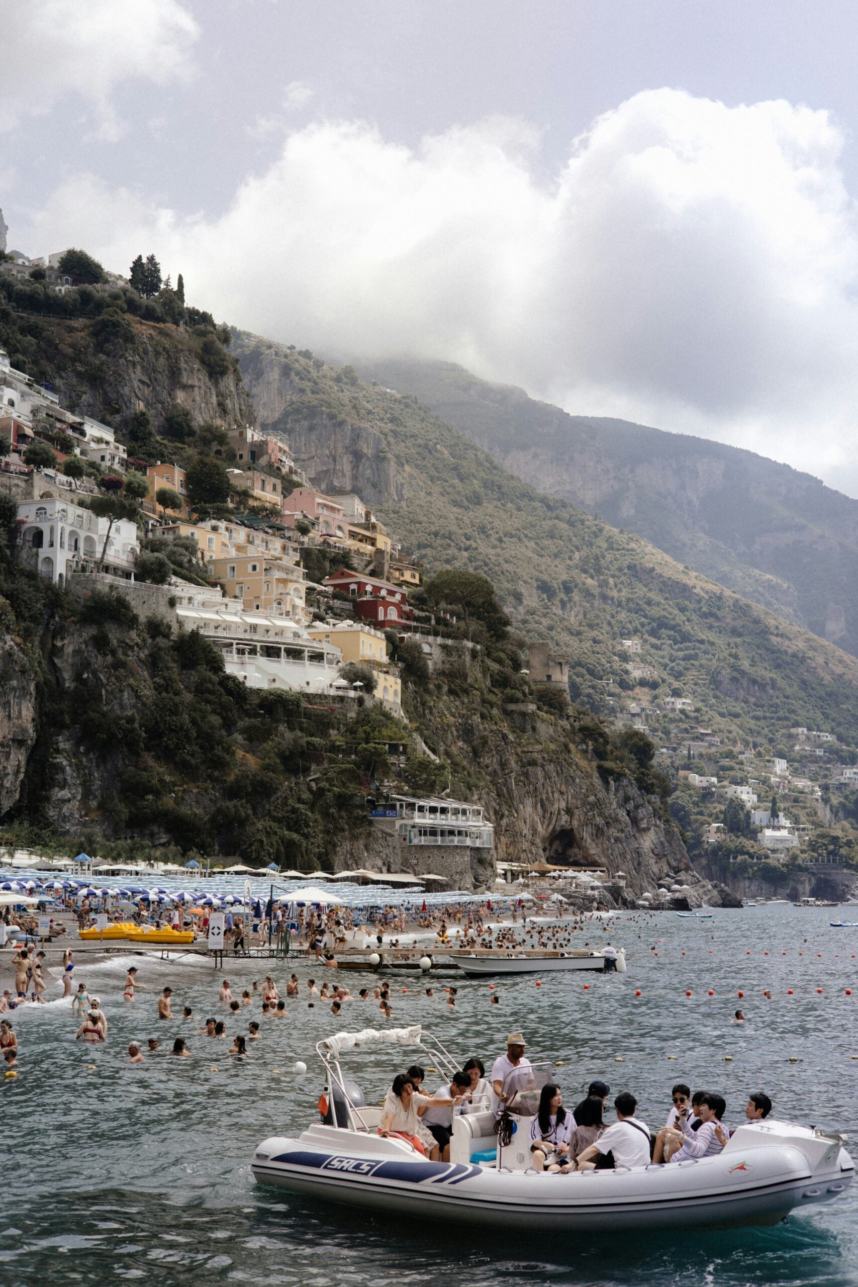 A crowded beach in Positano, Italy, bustling with tourists enjoying a sunny summer day. The beach is packed with colorful umbrellas, sunbathers, and swimmers, all relishing the warm Mediterranean sun and the crystal-clear waters of the Tyrrhenian Sea. The iconic, steep hillside of Positano, dotted with pastel-colored buildings, rises dramatically in the background, adding to the vibrant atmosphere. The lively scene captures the essence of Positano as a popular destination, where tourists flock to experience the stunning coastal views, refreshing sea, and the unique charm of this famous Amalfi Coast village.