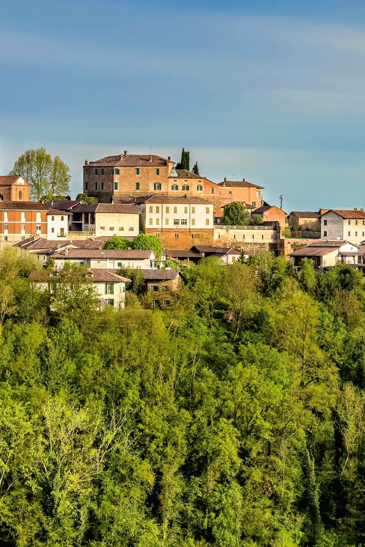A hilltop village with rustic buildings and red-tiled roofs is surrounded by lush green trees under a partly cloudy sky—the perfect scene to experience during the best time to visit Piedmont, Italy.
