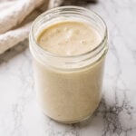 A glass jar filled with creamy, beige sourdough starter recipe sits on a white marble surface, complemented by a light-colored cloth in the background.