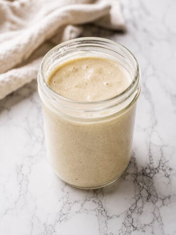 A glass jar filled with creamy, beige sourdough starter recipe sits on a white marble surface, complemented by a light-colored cloth in the background.