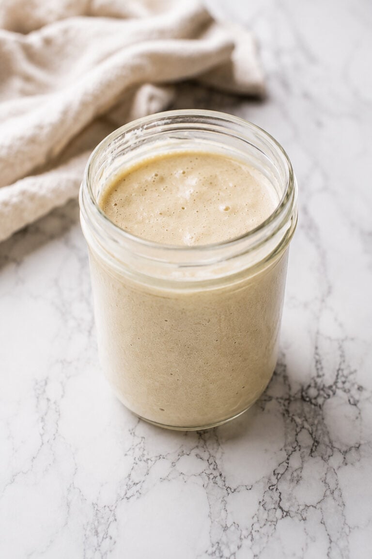A glass jar filled with creamy, beige sourdough starter recipe sits on a white marble surface, complemented by a light-colored cloth in the background.