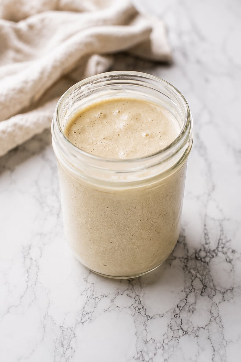 A glass jar filled with creamy, beige sourdough starter recipe sits on a white marble surface, complemented by a light-colored cloth in the background.