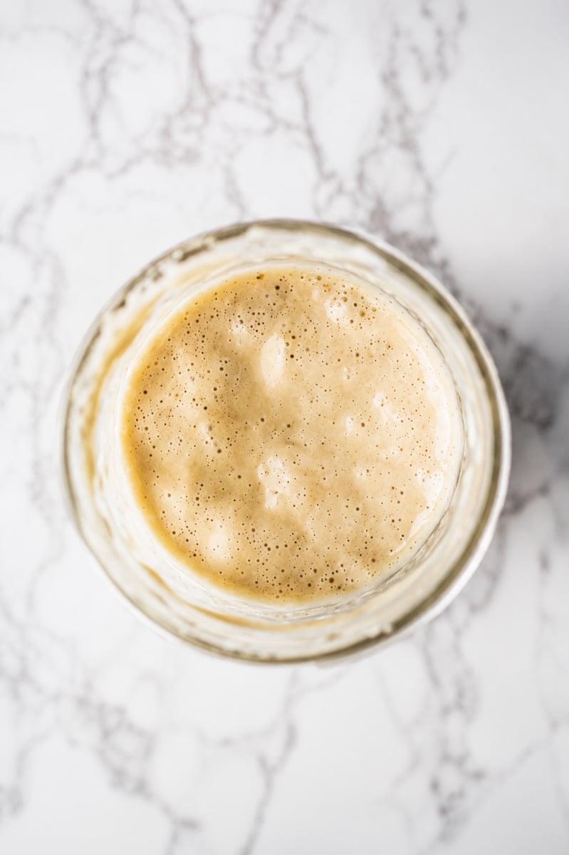 A close-up top view of an active sourdough starter in a glass jar, perfect for any sourdough starter recipe, showing a bubbly, frothy surface on a white marble background.