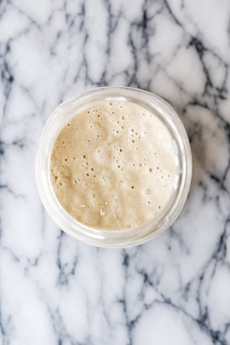 A glass jar filled with bubbly sourdough starter, perfect for learning how to revive sourdough starter, sits on a white marble surface, viewed from above.