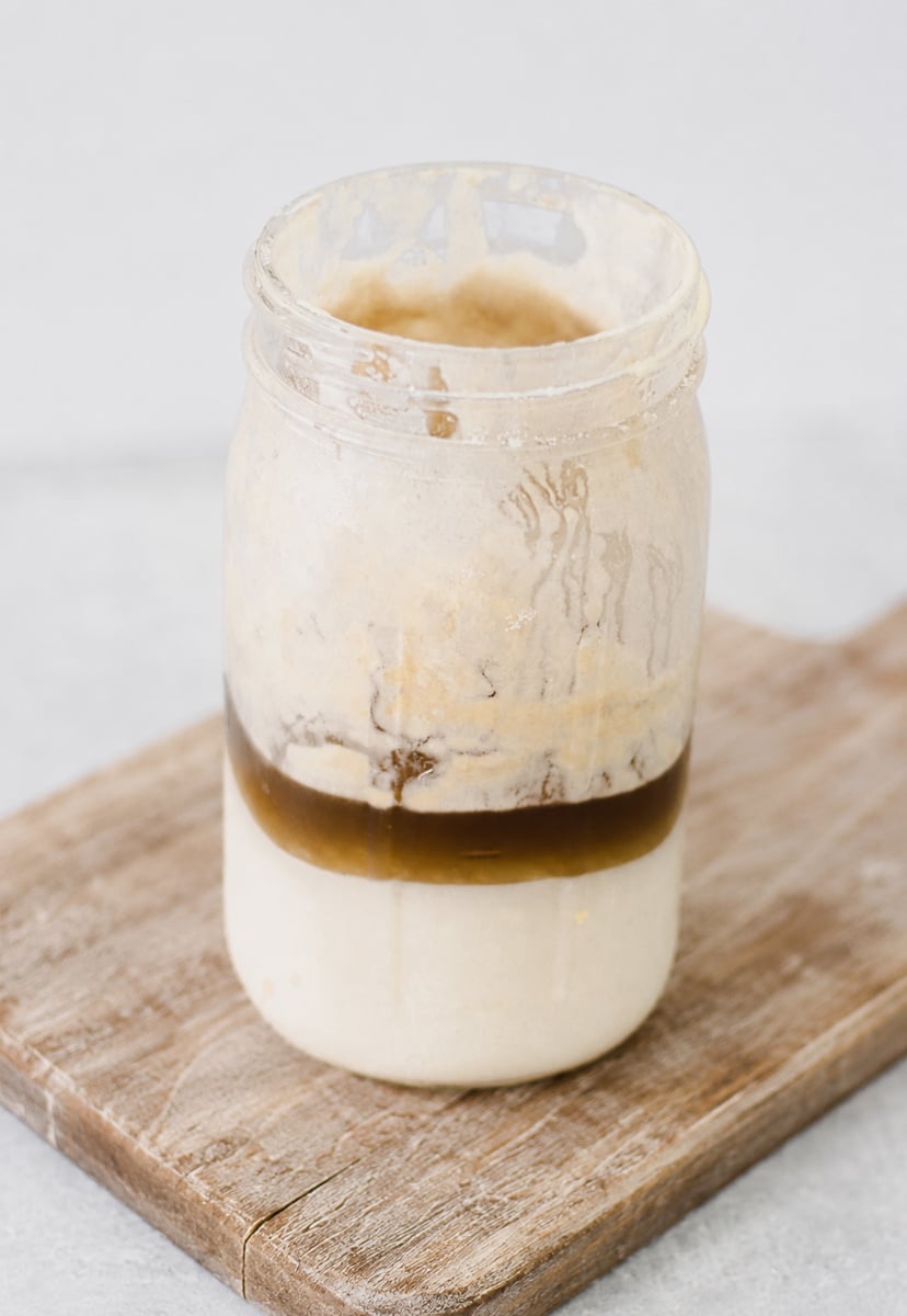 A glass jar with separated sourdough starter and dark hooch liquid on top sits on a wooden board, against a light gray background—a common sight when learning how to revive sourdough starter.