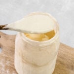 A wooden spoon holding creamy sourdough starter above a glass jar, which sits on a wooden cutting board with some flour scattered nearby-perfect for demonstrating how to revive sourdough starter at home.