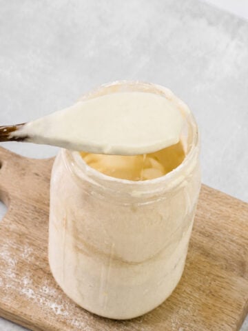 A wooden spoon holding creamy sourdough starter above a glass jar, which sits on a wooden cutting board with some flour scattered nearby-perfect for demonstrating how to revive sourdough starter at home.