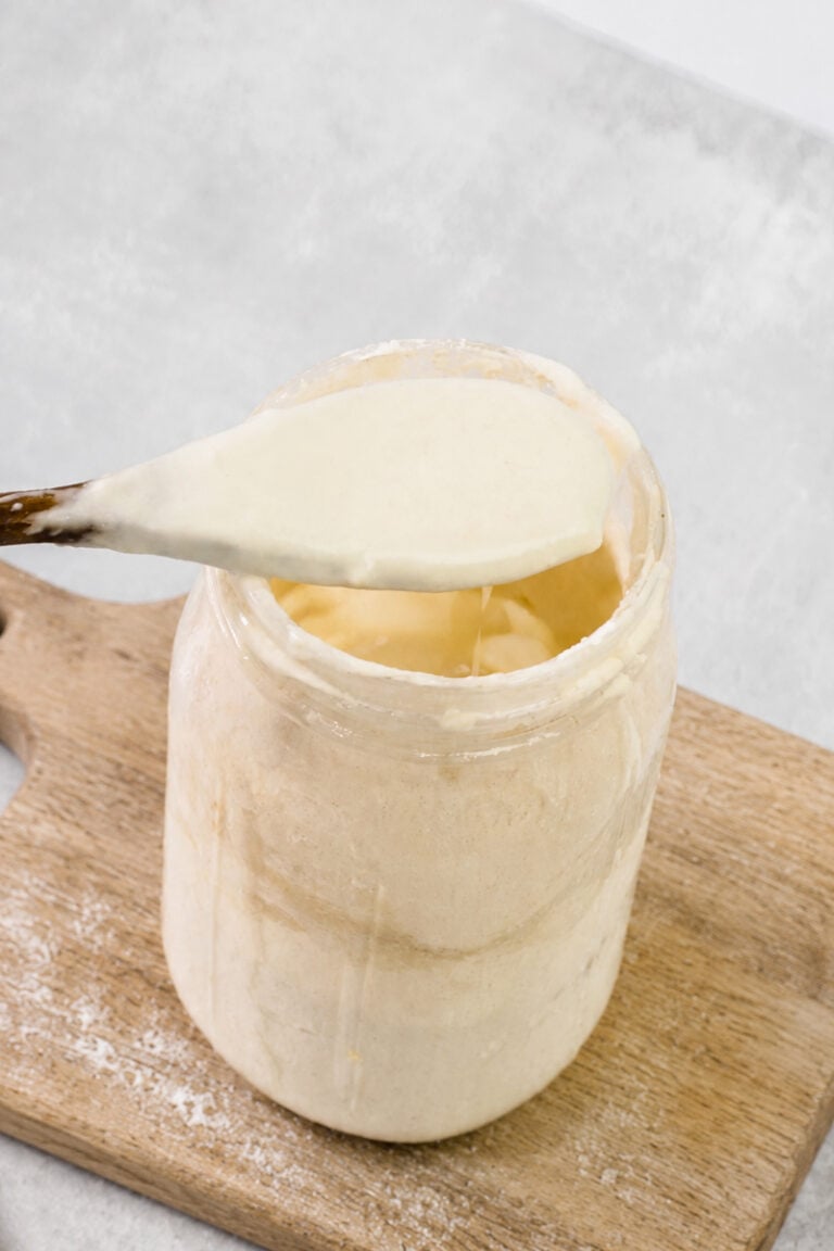A wooden spoon holding creamy sourdough starter above a glass jar, which sits on a wooden cutting board with some flour scattered nearby—perfect for demonstrating how to revive sourdough starter at home.