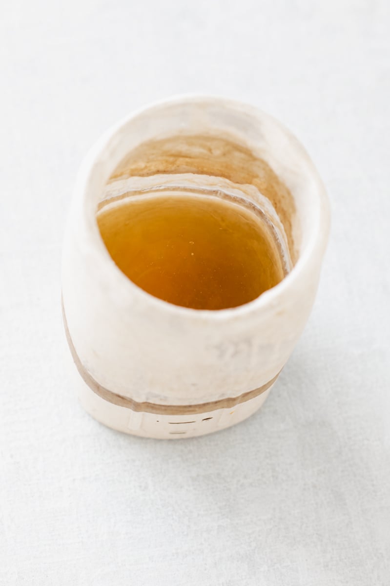 A close-up view of a handmade ceramic cup filled with a light brown liquid, possibly tea or broth, placed on a light, textured surface-evoking the calming ritual often enjoyed while learning how to revive sourdough starter.