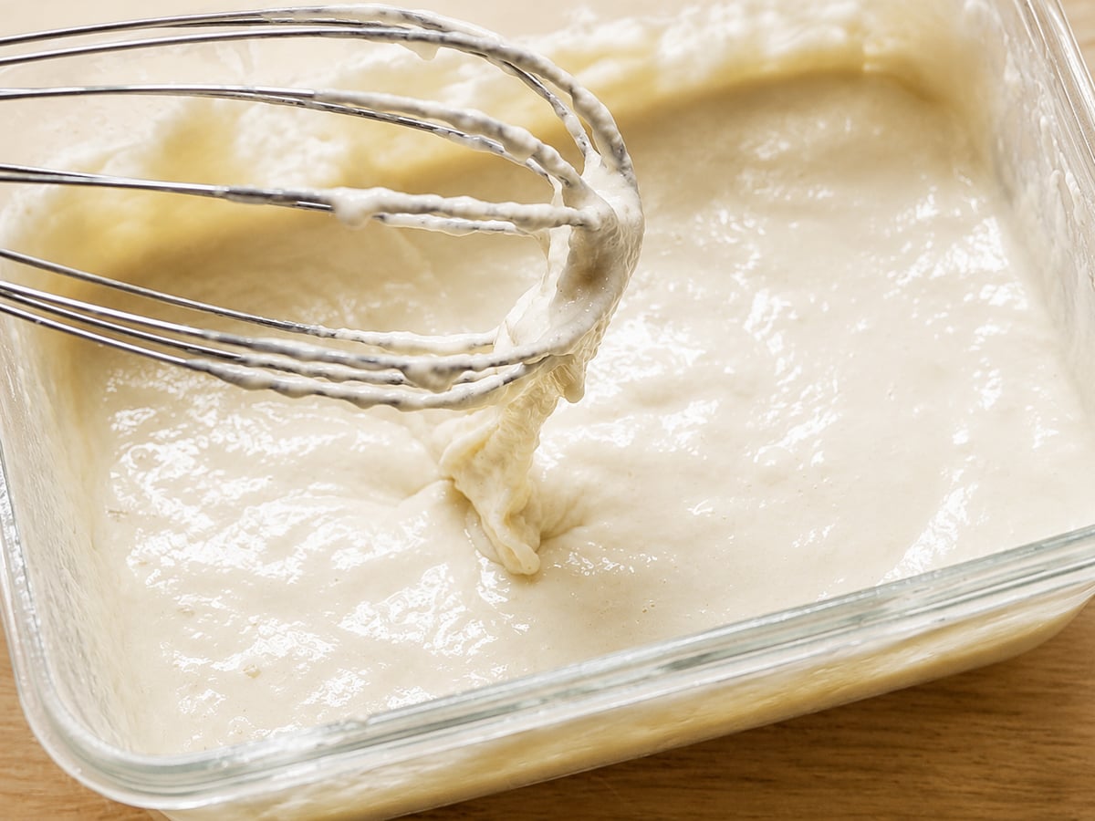 A close-up of a glass dish filled with thick, creamy batter being mixed with a metal whisk, showing the smooth texture—perfect for beginning your homemade sourdough starter recipe.