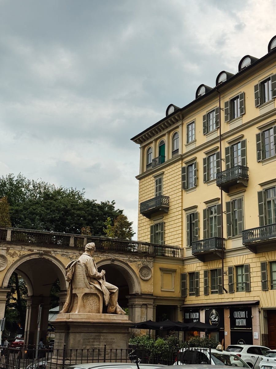 A stone statue of a seated figure sits in front of a yellow building with green shutters and balconies in Turin, Italy, beneath a cloudy sky. An arched walkway and some trees are visible in the background.