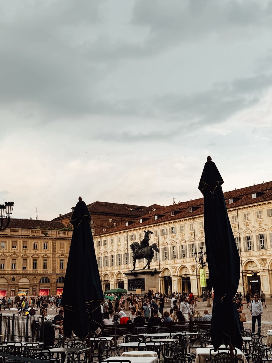 A lively European square in Turin, Italy, with people walking and gathering, surrounded by historic buildings. Two tall closed umbrellas and empty tables are in the foreground. A statue of a horse and rider stands in the center under a cloudy sky.