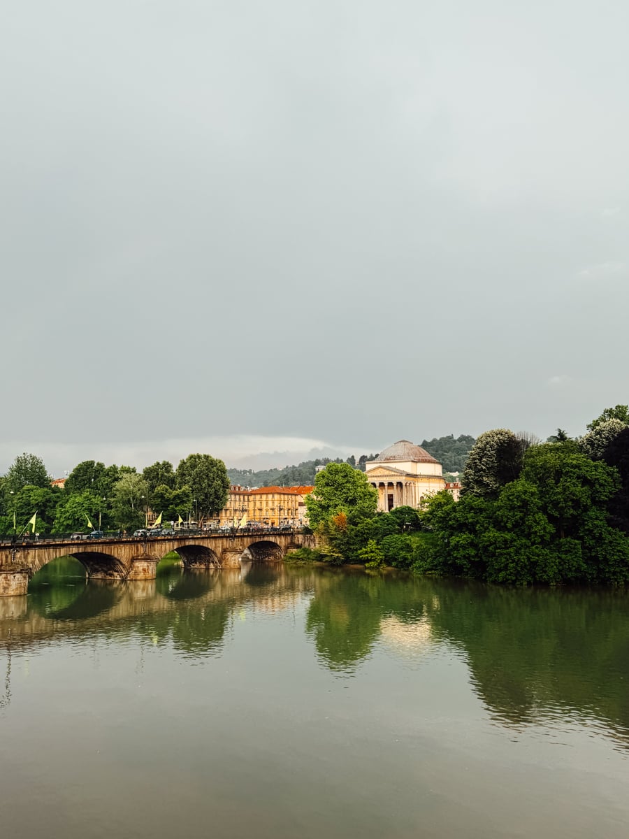 A stone bridge crosses a calm river in Turin, Italy, with trees, colorful buildings, and a domed classical structure in the background under a cloudy sky. The picturesque scene is beautifully reflected in the water.