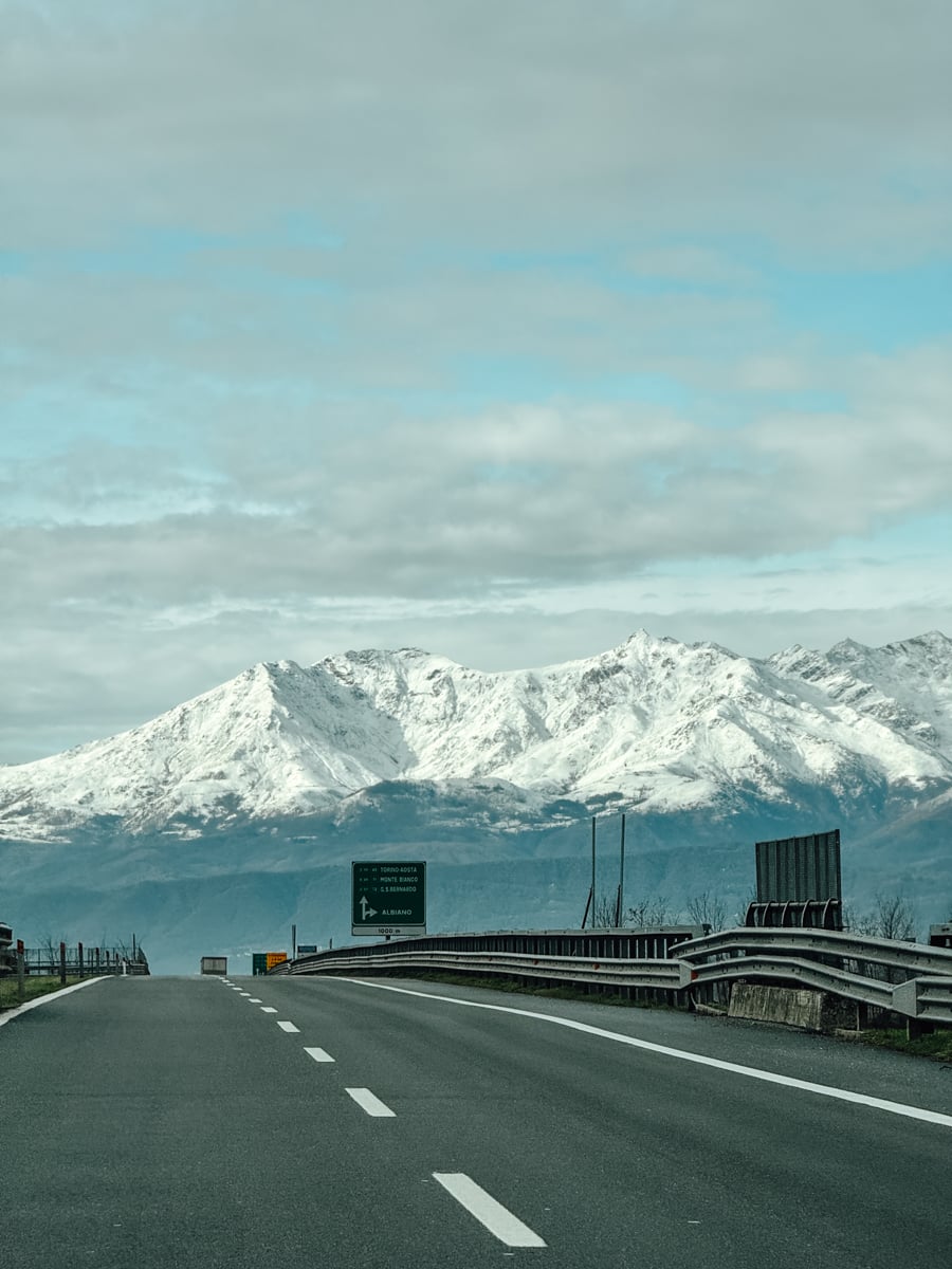 A paved road leads toward snow-capped mountains under a mostly cloudy sky near Turin, Italy, with road signs visible along the roadside and guardrails flanking the highway.