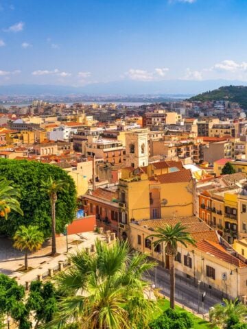 A scenic view of Cagliari, Sardinia-showcasing why Cagliari is famous-with colorful buildings, a church tower, lush green palm trees in the foreground, and the coastline visible under a radiant blue sky.