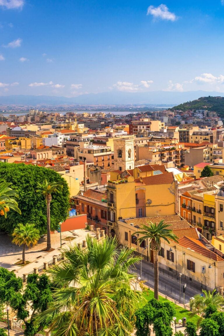 A scenic view of Cagliari, Sardinia—showcasing why Cagliari is famous—with colorful buildings, a church tower, lush green palm trees in the foreground, and the coastline visible under a radiant blue sky.