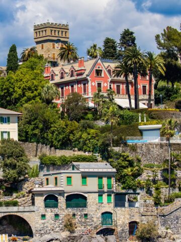 A large, elegant red villa with a tower sits on a lush hillside above stone buildings by the water-an idyllic scene that captures what Santa Margherita is known for: luxurious architecture, palm trees, and Mediterranean charm under a partly cloudy sky.