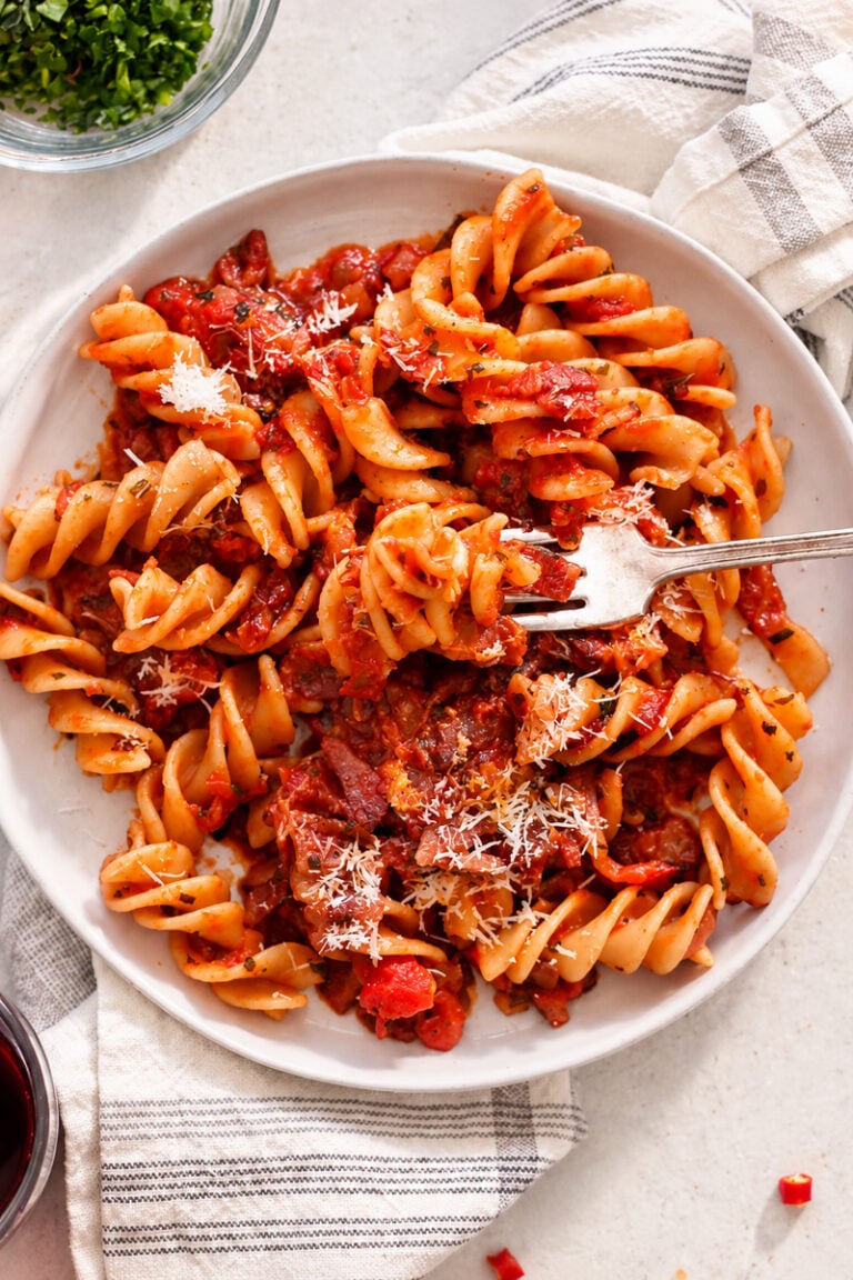 A bowl of fusilli amatriciana in a tomato-based sauce with bits of meat and a dusting of grated cheese, served with a fork.