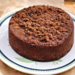 A round Chocolate Rye Crumb Cake sits on a decorative plate with a green pattern, placed on brown parchment paper atop a light-colored countertop. The cake has a crumbly, textured top.