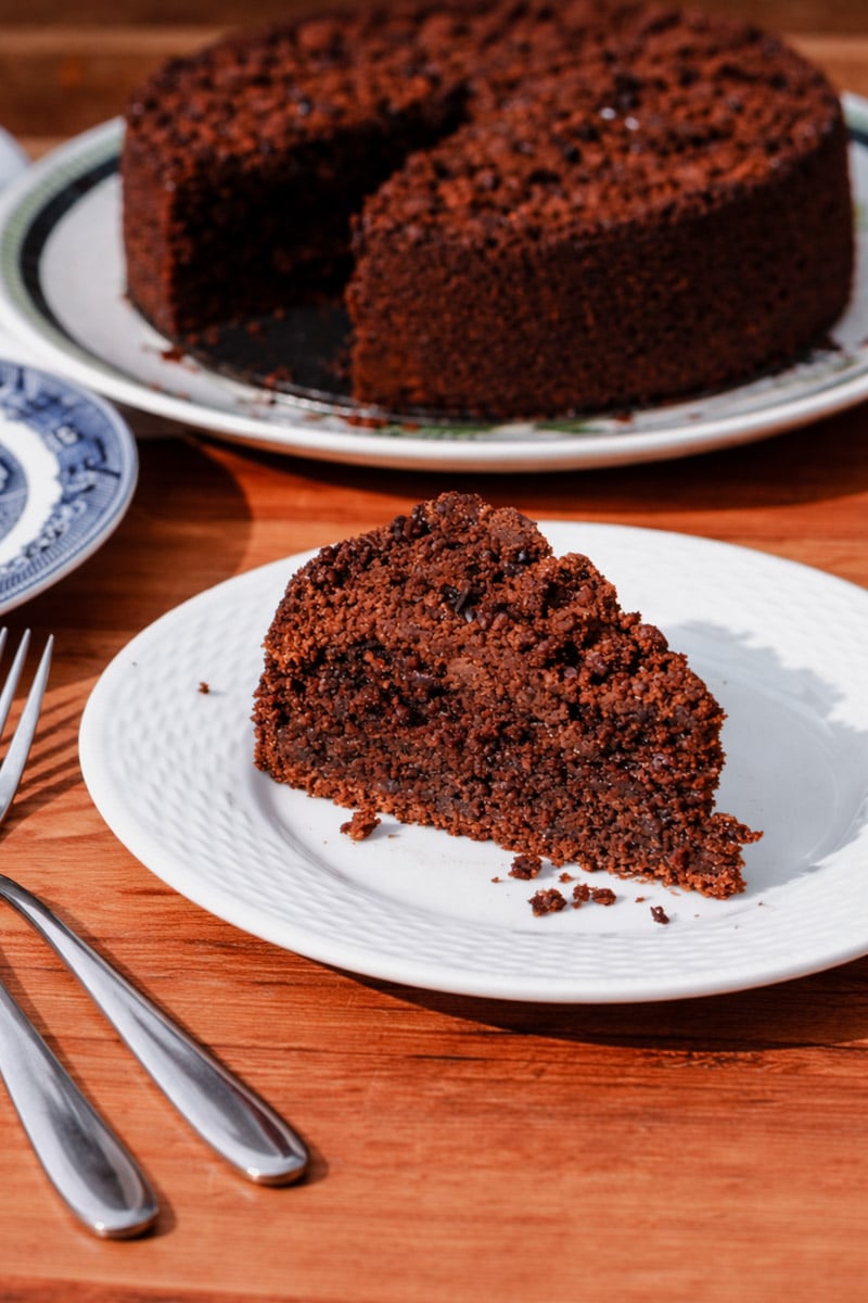 A slice of Chocolate Rye Crumb Cake on a white plate sits beside two forks on a wooden table, with the rest of the cake, missing one slice, visible in the background on another plate.