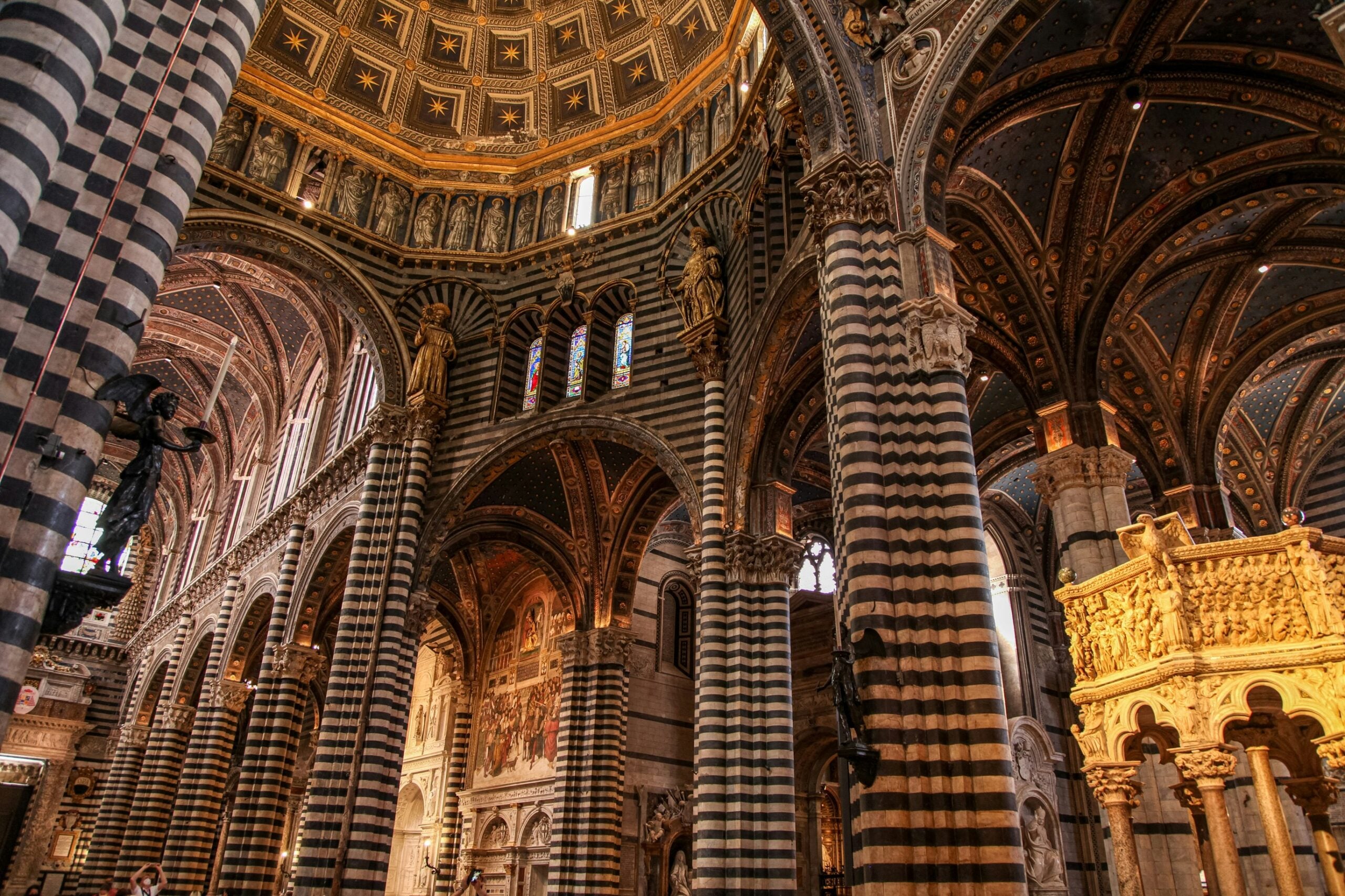 A stunning view inside the Duomo of Siena, Italy, showcasing its incredibly ornate interior. The photograph captures the magnificent striped marble columns, intricate mosaic floors, and lavishly decorated ceilings adorned with frescoes and gold accents. The grandeur of the high altar, detailed sculptures, and beautifully stained glass windows highlight the cathedral's exquisite craftsmanship and artistic splendor. This awe-inspiring scene reflects the rich cultural and religious heritage of Siena, Italy, making it a must-see for visitors.