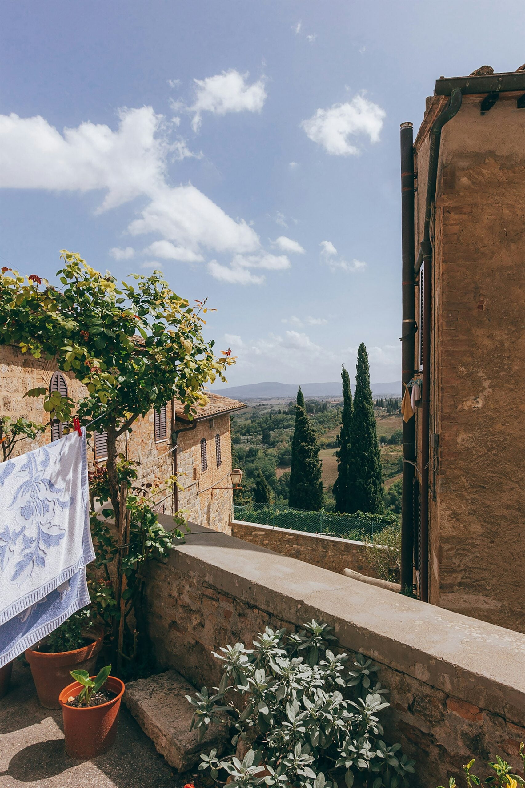 A stunning rooftop view of the Tuscan landscape, capturing the essence of this picturesque region. The photograph showcases terracotta rooftops of historic buildings, gently sloping into the lush, rolling hills covered in vineyards, olive groves, and cypress trees. The horizon is dotted with quaint farmhouses and winding country roads under a clear blue sky. This image epitomizes the serene and timeless beauty of Tuscany, offering a breathtaking perspective of the countryside from a vantage point in Siena, Italy.