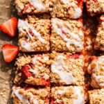 Strawberry Crumble Bars cut into squares, topped with a white icing drizzle, are arranged on parchment paper with fresh strawberry halves on the side for a deliciously sweet treat.