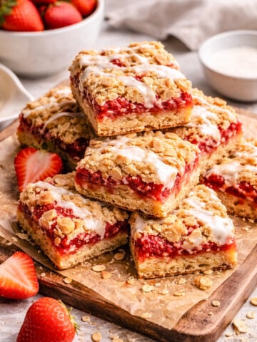 A stack of Strawberry Crumble Bars with a crumbly topping and white icing drizzle sits on parchment paper, surrounded by fresh strawberries and bowls of ingredients in the background.