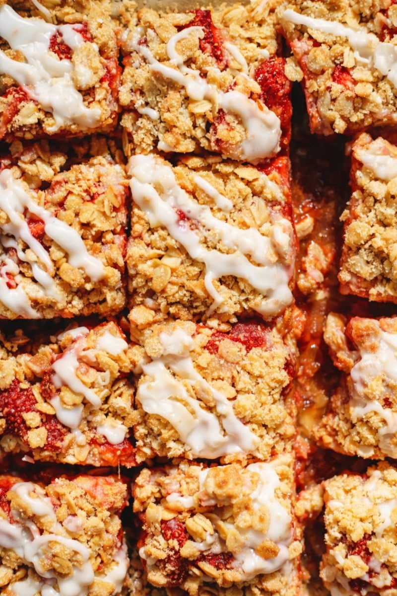 Close-up of several Strawberry Crumble Bars with a red fruit filling, drizzled with white icing, arranged closely together in a grid pattern.
