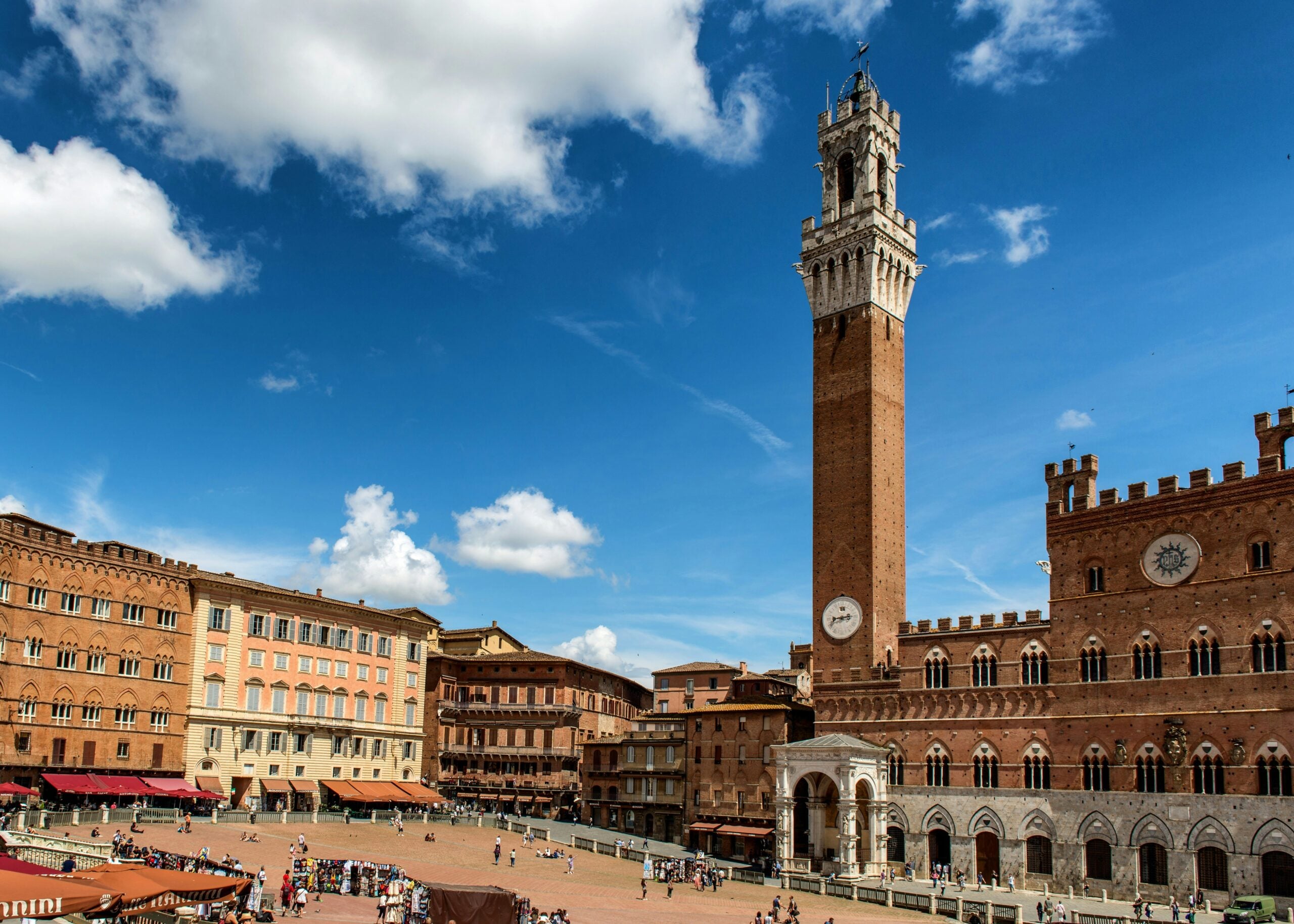 A vibrant photograph of the main square in Siena, Italy, known as Piazza del Campo. The image captures the unique fan-shaped layout, surrounded by historic medieval buildings and bustling with activity. The iconic Torre del Mangia stands tall in the background, adding to the charm and historic significance of the square. This picturesque scene highlights the cultural heart of Siena, Italy, and its architectural splendor.