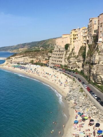 A scenic beach in Tropea, Italy, with turquoise water and golden sand is lined with colorful umbrellas and sunbathers. Tall buildings perch on cliffs overlooking the shoreline, with a winding road running along the edge.