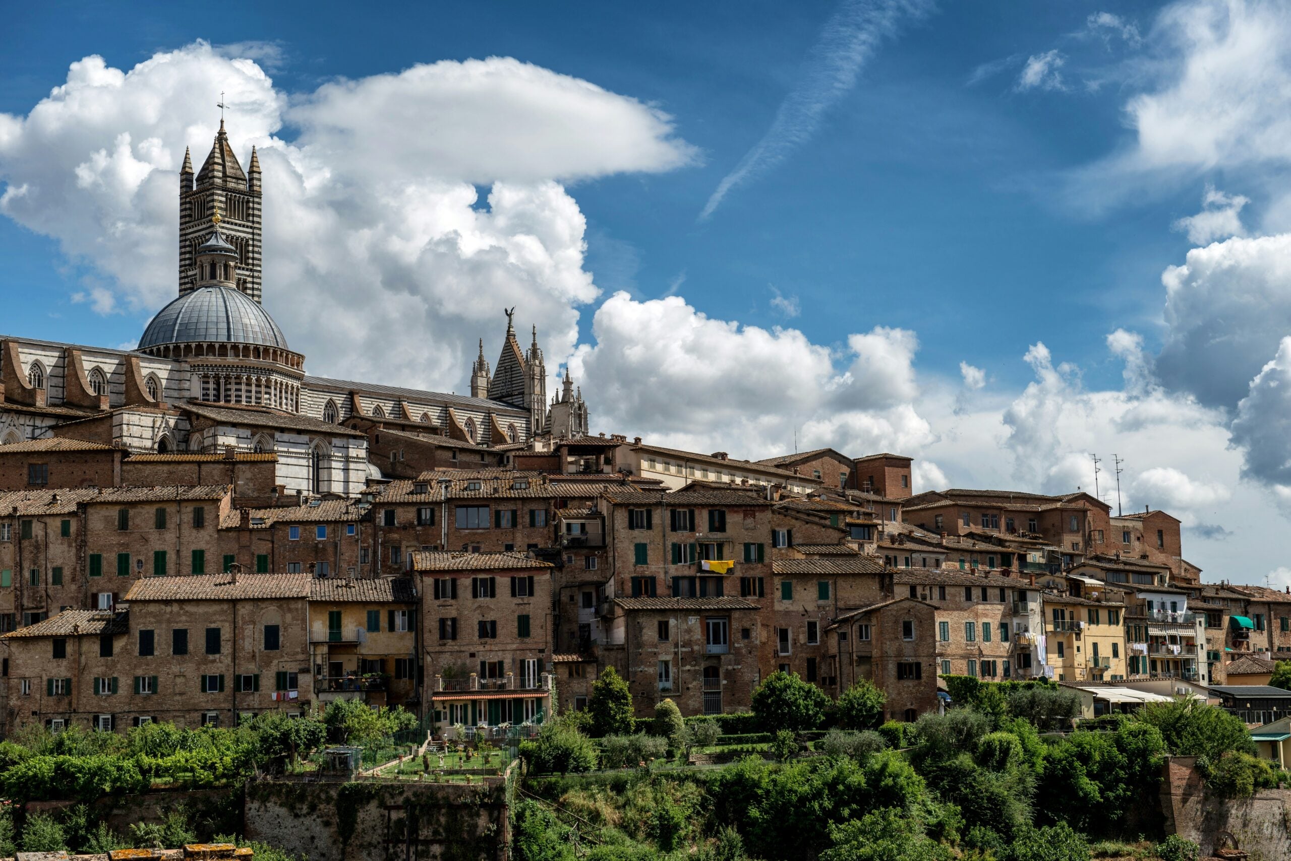 A breathtaking view looking up at the city of Siena, Italy, perched on a hill. The photograph captures the historic skyline with the prominent Torre del Mangia rising above the medieval buildings. The terracotta rooftops and ancient architecture showcase the timeless beauty and cultural significance of Siena, Italy, inviting travelers to explore this picturesque Tuscan city.