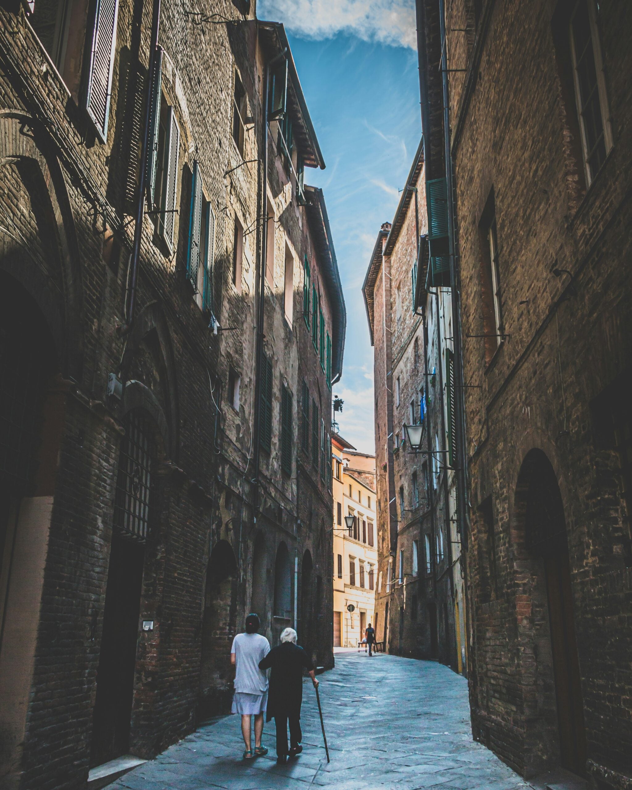 A heartwarming view of an old woman and her daughter walking along a narrow, cobblestone street in Siena, Italy. The ancient buildings with their rustic facades and shuttered windows line the path, creating a charming and timeless atmosphere. The pair's leisurely stroll through this historic street captures the essence of Siena's rich cultural heritage and the beauty of everyday life in this medieval city.