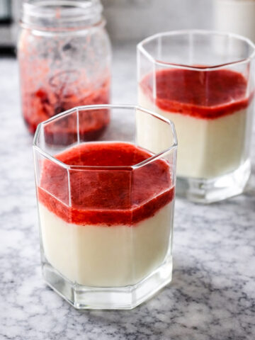 Two clear glasses filled with strawberry panna cotta are topped with bright red strawberry sauce and sit on a marble countertop. A jar with more strawberry sauce is in the background.