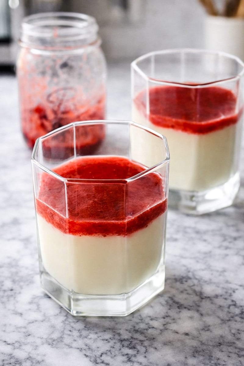 Two clear glasses filled with strawberry panna cotta are topped with bright red strawberry sauce and sit on a marble countertop. A jar with more strawberry sauce is in the background.