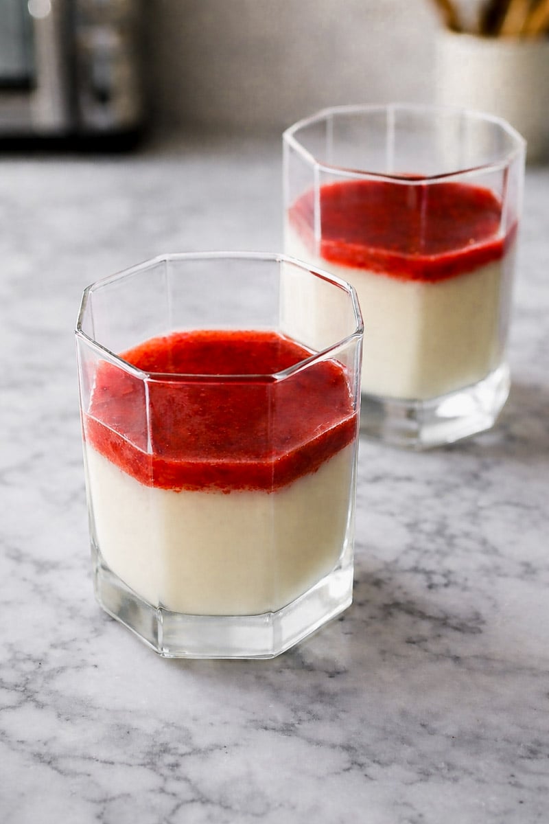 Two clear, faceted glasses filled with creamy white strawberry panna cotta topped with a bright red sauce, set on a gray marble surface. The background is softly blurred.