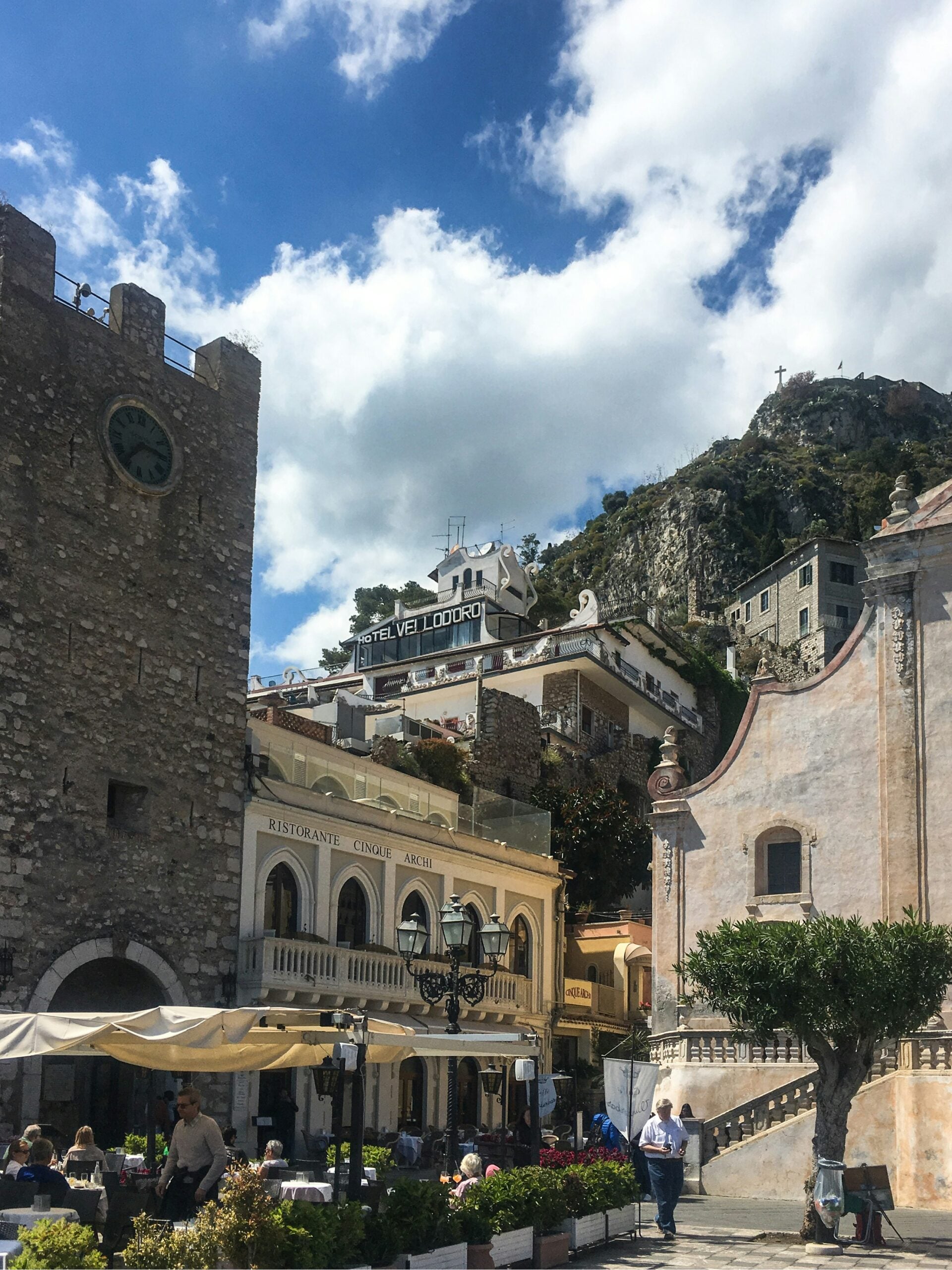 A quaint small piazza in Taormina, Sicily, exudes charm with its cobblestone pavement, surrounded by historic buildings adorned with vibrant flowers. The centerpiece of the piazza is a beautiful fountain, where locals and tourists gather to enjoy the serene atmosphere. Outdoor cafes with inviting seating areas spill onto the square, offering a perfect spot to savor Sicilian cuisine while soaking in the picturesque surroundings. This charming piazza captures the essence of Taormina's warm and welcoming spirit.