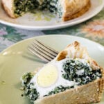 A slice of savory torta pasqualina with spinach and a hard-boiled egg inside sits on a green plate next to a fork. In the background, the rest of the pie is on a floral-patterned plate.