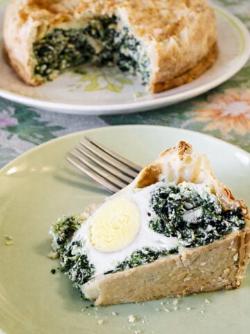 A slice of savory torta pasqualina with spinach and a hard-boiled egg inside sits on a green plate next to a fork. In the background, the rest of the pie is on a floral-patterned plate.