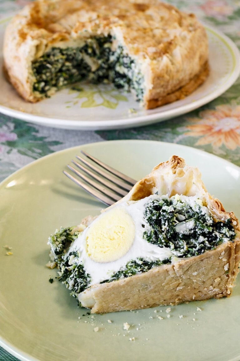 A slice of savory torta pasqualina with spinach and a hard-boiled egg inside sits on a green plate next to a fork. In the background, the rest of the pie is on a floral-patterned plate.