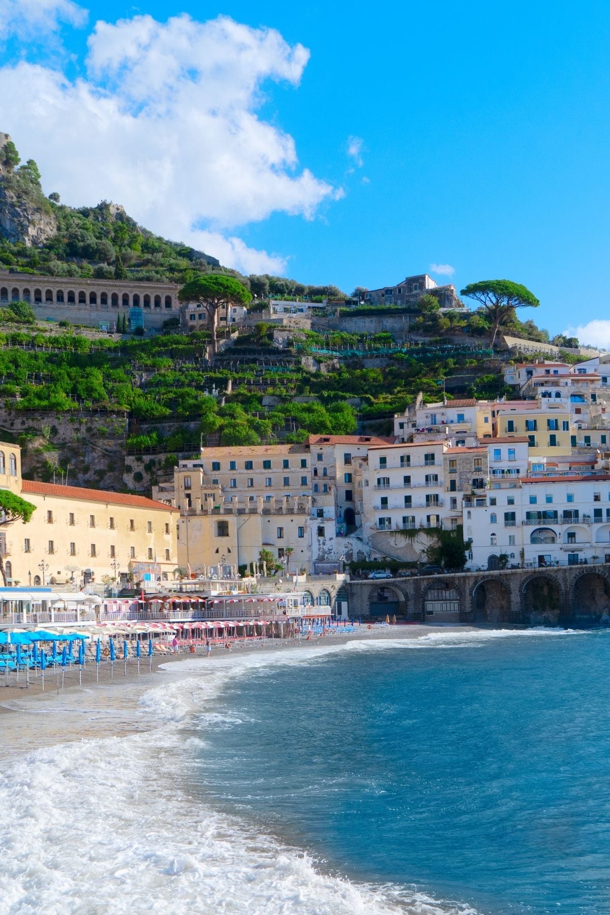 Colorful buildings and terraced hills line a sunny coastline, showcasing the charm of Amalfi Coast towns with clear blue water and a sandy beach filled with umbrellas and chairs under a bright blue sky with scattered clouds.