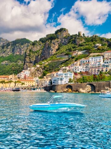 A small white speedboat floats on bright blue water near vibrant Amalfi Coast towns, with colorful buildings, green hills, and rocky cliffs under a partly cloudy sky.