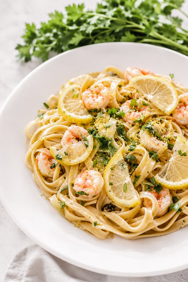 A white plate of fettuccine pasta topped with succulent shrimp, lemon slices, parsley, and grated lemon zest. This vibrant dish is the perfect example of Easy Shrimp Scampi with Linguine, with fresh parsley visible in the background to complete the inviting presentation.