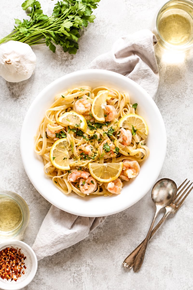 A white plate of Easy Shrimp Scampi with Linguine is served with lemon slices and garnished with fresh herbs. Surrounding the plate are fresh parsley, garlic, a napkin, wine glasses, utensils, and a small bowl of red pepper flakes.